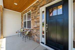 A front porch with a black door, stone and siding exterior walls, two metal chairs with yellow cushions, and a concrete floor. A window is next to the door, and the ceiling is wood-paneled.