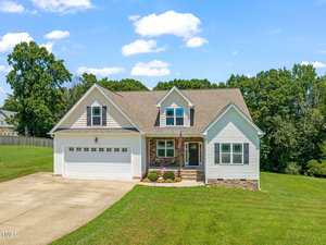 A two-story suburban house with beige siding, stone accents, a gable roof, a front porch, double garage, and a well-maintained lawn, set against a backdrop of trees under a blue sky with scattered clouds.