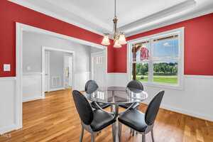 A dining area with red and white walls, a glass-top round table, and four black chairs. A window shows a green lawn and American flag outside. Hardwood floors and a ceiling light fixture are visible.