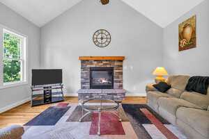 A living room with a vaulted ceiling features a stone fireplace, a wall clock above it, a TV on a stand, a beige sofa with a lamp, a glass coffee table, and a multicolored rug on a wooden floor.