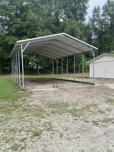 A metal carport with a peaked roof and open sides stands on a gravel area next to a light-colored building, surrounded by grass and trees.