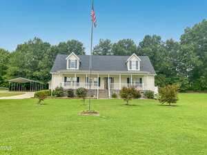 A two-story house at 920 Vanco Mill Road in Henderson features light-colored siding, a dark roof, and dormer windows. It sits on a large, well-maintained lawn with small shrubs, an American flag out front, and trees lining the background.