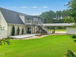 A two-story beige house with a covered second-floor balcony, large wooden deck, outdoor bar area, detached garage, and green lawn bordered by trees on a clear day.