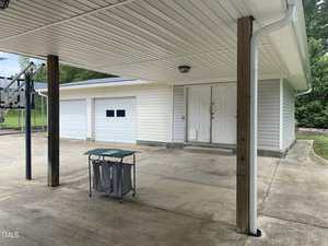 Covered concrete driveway with wooden posts, a basketball hoop on the left, a laundry hamper in the center, and a white garage with two doors and a double entry door in the background. Trees are visible in the background.