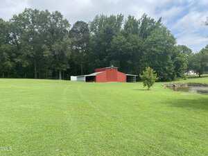 A red barn with a white roof sits on a large, grassy lawn. Trees line the background, and there is a small pond to the right of the barn. The sky is partly cloudy.