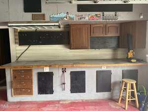 A workshop with a wooden workbench, drawers, brown cabinets, pegboards, a yellow power tool, a wooden stool, and various signs and decorations on the upper wall. The floor is red and the space appears organized.