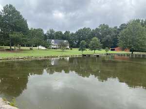 A pond with still water reflecting trees and a cloudy sky, with a grassy lawn, a white house, and a red barn-like building in the background. A small dock extends over the water near the trees.
