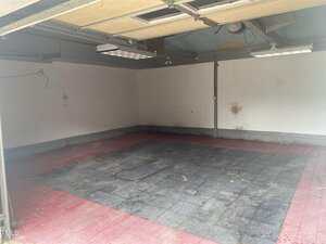 An empty garage with exposed beams, white walls, two open garage doors, and a floor with red and black tiles. There are some stains and marks on the walls and floor.