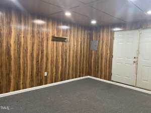 A small room with wood-paneled walls, gray carpet, a small wall shelf, an electrical panel, and double white doors. The ceiling has recessed lighting.
