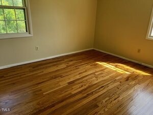 Empty room with wood flooring, cream-colored walls, and two windows letting in natural light. Sunlight forms a bright patch on the floor near one window. No furniture or decorations present.