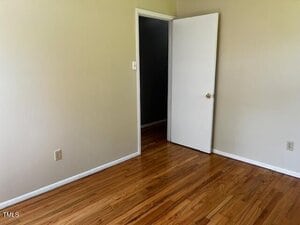 Empty room with light-colored walls, wood flooring, an open white door, electrical outlet, and baseboard visible. The door leads to a darker hallway with the same wood flooring.