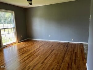 An empty room with hardwood floors, gray walls, a white ceiling, and a ceiling fan. Large window on the left lets in natural light. Electrical outlets are visible on the walls.