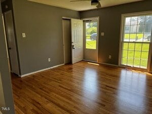 A room with light-colored hardwood floors, gray walls, a white door, a ceiling fan, and large windows letting in sunlight. The view outside shows green grass and a road with parked cars.