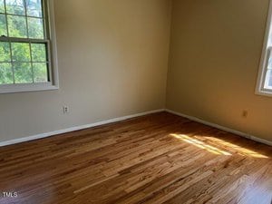 Empty room with light-colored walls, hardwood flooring, and two windows letting in natural light. Sunlight creates a bright patch on the floor near the corner.