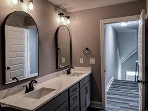 A modern bathroom features a double vanity with two rectangular sinks, black fixtures, two large arched mirrors, and light gray cabinets. The room opens to a hallway with wood-like flooring and a staircase.