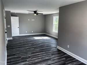 A spacious, empty room with gray walls, white trim, dark wood-look flooring, a ceiling fan with light, a white door, and a large window letting in natural light.