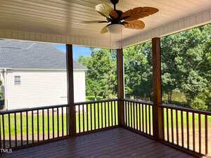 A screened-in porch with wooden floors and railings, a ceiling fan with leaf-shaped blades, and a view of a grassy backyard with trees and a neighboring white house.
