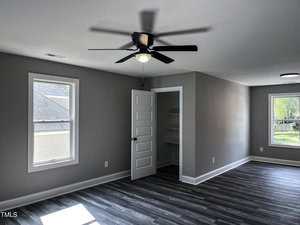 A room with gray walls, dark wood flooring, a ceiling fan with a light, two windows, an open closet with shelves, and an open door letting in natural light.