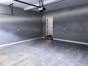 An empty garage with gray walls, a concrete floor, a white door with steps leading up to it, and a ceiling-mounted garage door opener. There are no vehicles or items visible in the space.