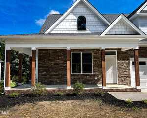 A single-story house features a covered front porch with wooden posts, a white door, stone facade, and white siding under a gray shingled roof. There are small shrubs in the front yard.