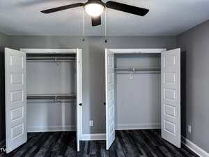 A room with two open closets side by side, each with a hanging rod, white double doors, gray walls, dark wood flooring, and a black ceiling fan with a light.