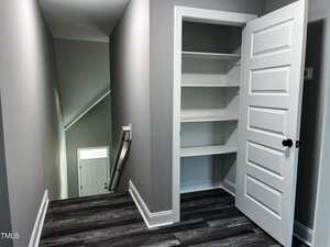 A hallway with dark wood flooring, gray walls, and white trim features an open door revealing a closet with four shelves. A staircase with a white railing leads down to a door with a frosted glass window.