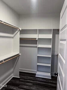 A walk-in closet with white walls, multiple wooden hanging rods, white shelving units, and dark wood flooring. The door is open, revealing the organized storage space.