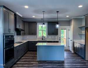 Modern kitchen with dark gray cabinets, white countertops, center island with sink, built-in oven, cooktop, dishwasher, pendant lights, and large windows letting in natural light. Wood-look flooring and light gray walls.