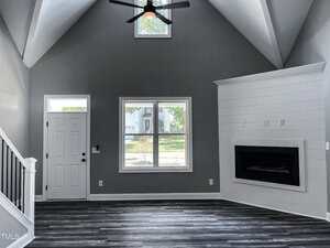 A living room with gray walls, vaulted ceiling, and wood-like flooring. Features a central window, a white front door to the left, and a modern black fireplace with white shiplap to the right. Ceiling fan overhead.