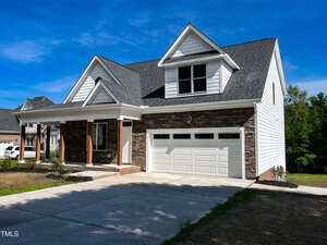 A two-story suburban house with a stone and white siding exterior, a covered front porch with wooden columns, and a two-car garage. The driveway and lawn are visible under a clear blue sky.