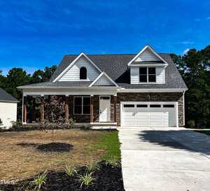 A two-story suburban house with white siding, brick accents, a covered front porch, and a double garage, sits on a lawn with young trees and landscaping, under a clear blue sky.