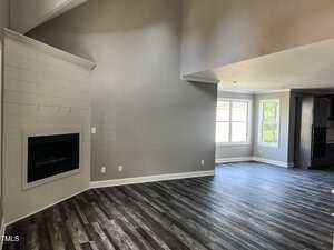 A modern, empty living room with gray walls, vaulted ceilings, a corner fireplace, large windows, and wood-look vinyl flooring extending into a dining area near the windows.