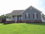 Charming single-story house at 2662 Walas Court in Creedmoor with gray siding, red shutters, and a brick foundation. Enjoy a front porch with white railings, arched windows, and a grassy lawn under a clear sky.