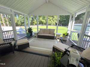 A screened-in porch with a vaulted ceiling, wicker furniture with beige cushions, a large rug, potted plants, and views of a grassy yard and trees through the windows.