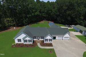 Aerial view of a single-story house at 5720 Raleigh Road, Kittrell, featuring a dark green roof, white exterior, three-car garage, curved walkway, manicured lawn, and surrounded by trees and outbuildings in the background.