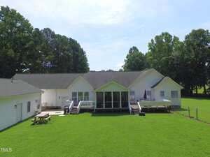 A large white single-story house with a screened porch, multiple decks, and a fenced backyard with green grass. There are trees in the background and a picnic table on the lawn.