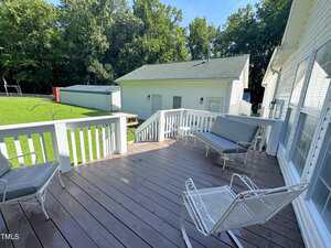 A wooden deck with white railings features two chairs and a bench with gray cushions. The deck overlooks a green lawn, a white garage, and several trees in the background.