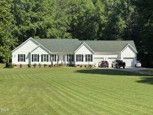 A single-story white house with a green roof, multiple windows, and a three-car garage sits in front of a large, neatly mowed lawn, surrounded by tall green trees. Two cars are parked in the driveway.