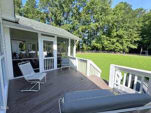 A wooden deck with white railings features patio chairs and a bench. The deck is attached to a screened porch and overlooks a large, grassy backyard bordered by trees and a wire fence.