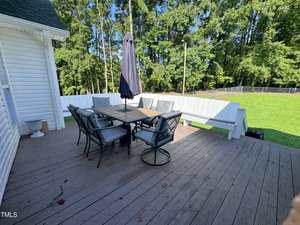 A wooden deck with a dining table, six chairs, an umbrella, and built-in white bench seating. The deck overlooks a grassy yard bordered by trees and a black fence.