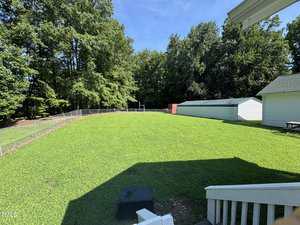 A fenced backyard with green grass, bordered by trees. There is a red shed and a long white storage building near the back, and part of a white house and porch are visible in the lower right corner.