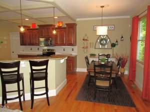 A kitchen and dining area with wooden cabinets, granite countertops, stainless steel appliances, pendant lighting, and a dining table with six chairs on a dark rug near a window.