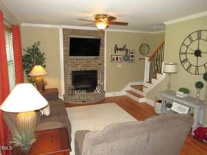A living room with a sofa, two armchairs, and two lamps. A brick fireplace with a TV above it is centered on the wall. A staircase with a white railing is to the right. The decor includes plants, framed art, and a large wall clock.
