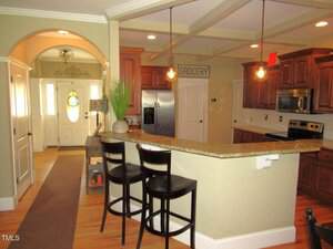 A kitchen with granite countertops, two black bar stools, stainless steel appliances, wood cabinets, and pendant lights. A decorative plant sits on the counter near an arched entryway and front door.