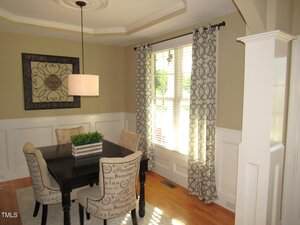A small dining area with a square dark wood table, four upholstered chairs, a decorative wall piece, a hanging light fixture, light beige walls, and two windows with patterned curtains letting in natural light.