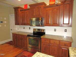A kitchen with wood cabinets, granite countertops, stainless steel oven and microwave, white tile backsplash, and hardwood floors. There are decorative items on top of the upper cabinets.