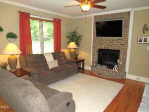 A living room with two brown reclining sofas, a brick fireplace with a mounted TV above it, two table lamps, a potted plant, wall decor, and a large window with brown curtains letting in natural light.