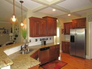 A kitchen with wooden cabinets, granite countertops, stainless steel dishwasher and refrigerator, pendant lights, a double sink, a coffee maker, and a red rug on a polished wood floor.