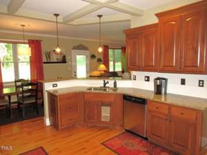 Kitchen with wooden cabinets, granite countertops, and stainless steel dishwasher. A coffee maker sits on the counter. The kitchen opens to a dining area with a table, red curtains, and windows letting in natural light.