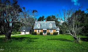 A two-story brick house with a steep roof and dormer windows sits on a large grassy lawn. Two leafless trees are in the foreground, with green bushes by the home's entrance and blue sky above.
